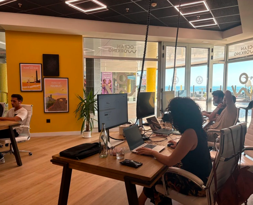 Teamworking in ufficio workation a Gran Canaria con persone al lavoro su laptop, vista sull'oceano attraverso grandi finestre.